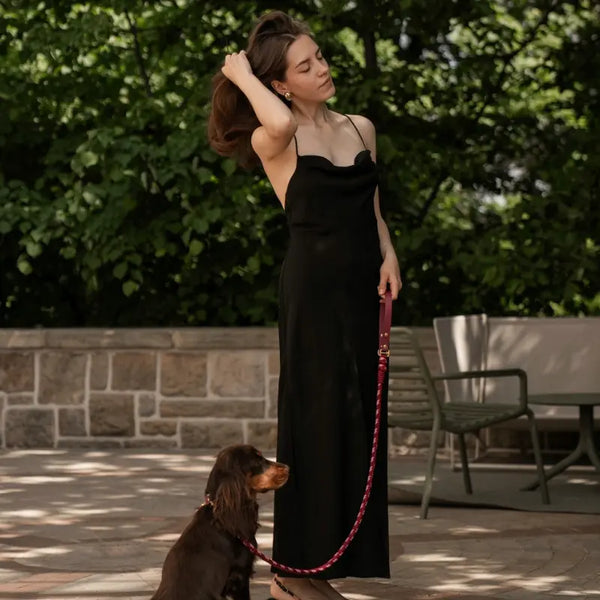 Woman in a black dress holding a dog leash with a dog sitting on the ground outdoors.